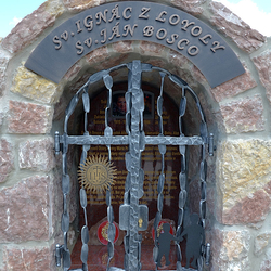 Kovaný pamätník svätých s atribútmi na mrežiach. Sv. Ignác z Loyoly - nápis IHS, Sv. Ján Bosco - deti